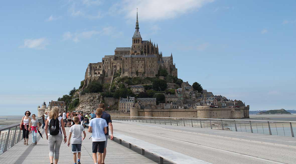 Man, woman, and boy walking along a footpath towards Mont Saint-Michel beneath a blue sky surrounded by golden sands